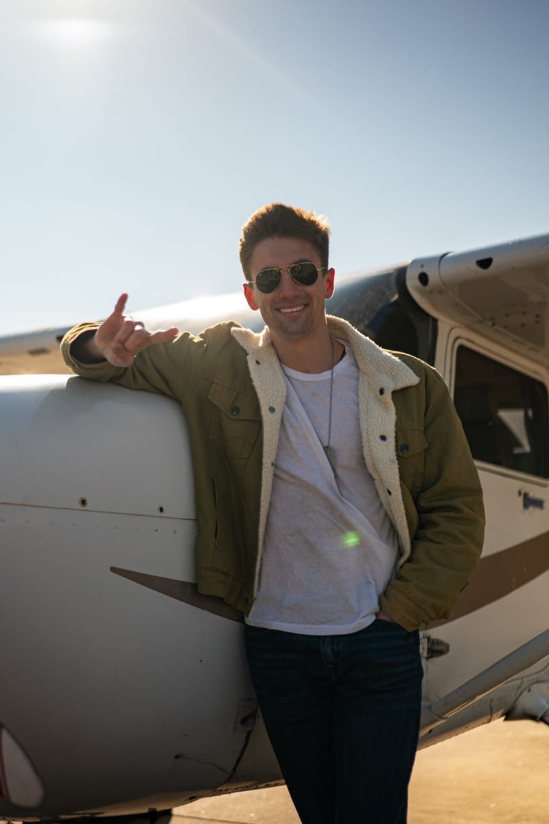 Armando leaning against a plane, throwing the Hook 'Em Horns sign with aviator sunglasses