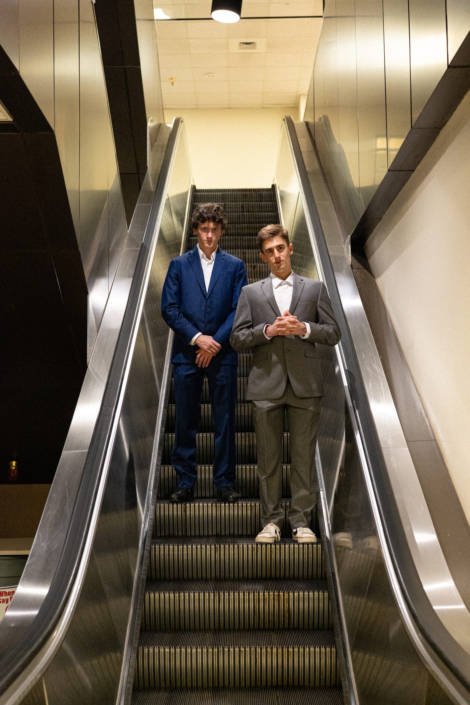 Armando and Ben descending an escalator in suits, dramatic low angle